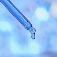 Kropla błękitnego serum z peptydami miedziowymi zwisa z pipety na rozmytym, niebieskim tle.