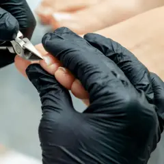 Dłoń w czarnej rękawiczce precyzyjnie wykonuje rekonstrukcję paznokcia u stopy za pomocą cążków.