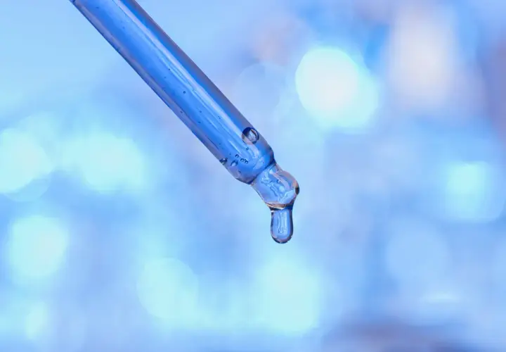 Kropla błękitnego serum z peptydami miedziowymi zwisa z pipety na rozmytym, niebieskim tle.