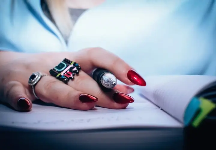 Dłoń z błyszczącymi, czerwonymi paznokciami i pierścionkami trzyma długopis nad notatnikiem. Dbałość o detale, jak witaminy na paznokcie, podkreśla styl.