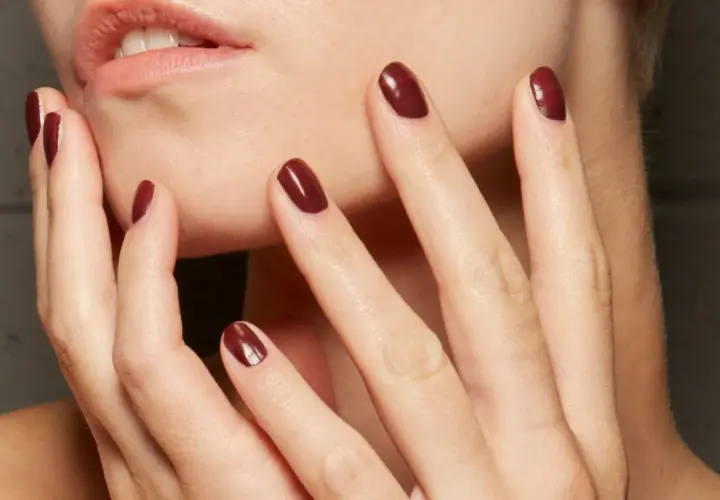 Dłonie z idealnym, ciemnoczerwonym manicurem kombinowanym. Delikatne muśnięcie policzka podkreśla elegancję.