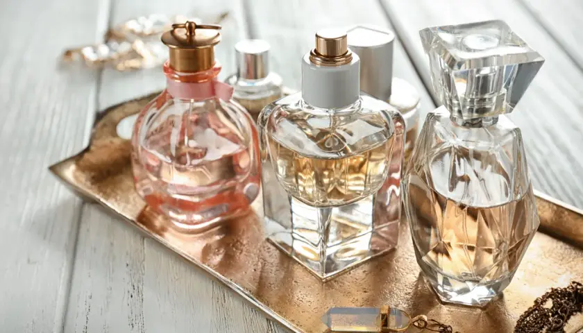 Eleganckie flakoniki perfum, w tym jeden z delikatną różową wstążką, stoją na złotej tacy. W tle błyszcząca biżuteria. Zapach, być może z nutą hexyl cinnamal, unosi się w powietrzu.