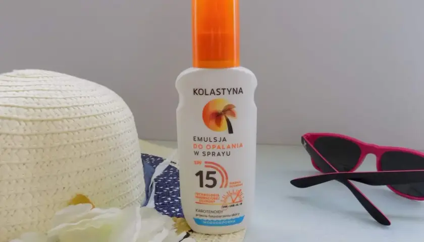 Emulsja do opalania w sprayu Kolastyna SPF 15 z karotenoidami, wodoodporna. Obok kapelusz i okulary przeciwsłoneczne.