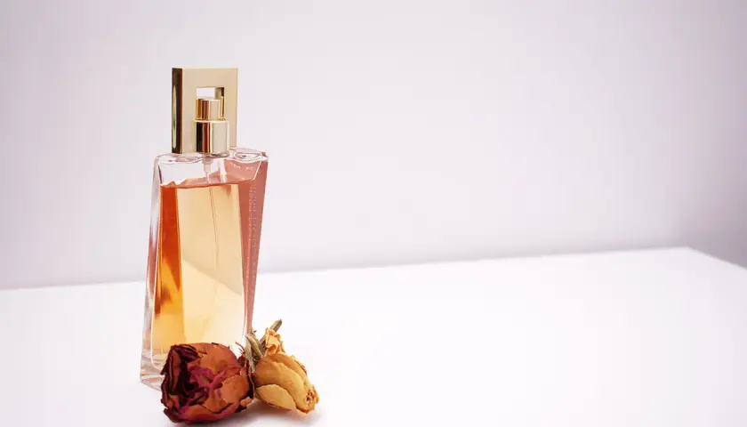 Elegancka butelka perfum z płynem w kolorze bursztynu, ozdobiona złotą nakrętką. Obok leżą dwie zasuszone róże, sugerujące nuty piżma w zapachu.