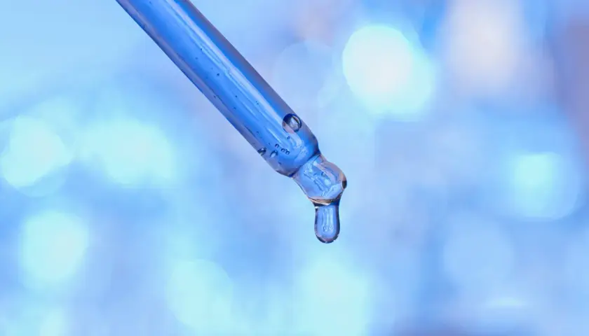 Kropla błękitnego serum z peptydami miedziowymi zwisa z pipety na rozmytym, niebieskim tle.