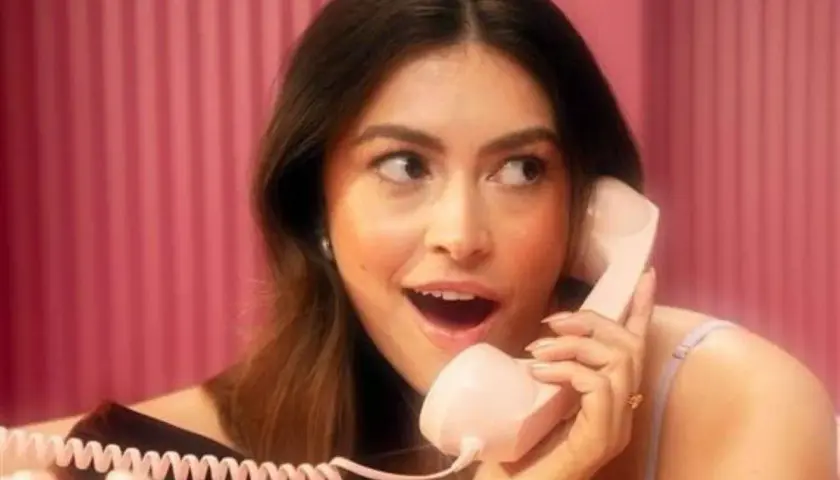 Młoda kobieta z zaciekawieniem rozmawia przez różowy telefon. Jej makijaż jest nienaganny, co może sugerować pozytywne mary kay opinie.