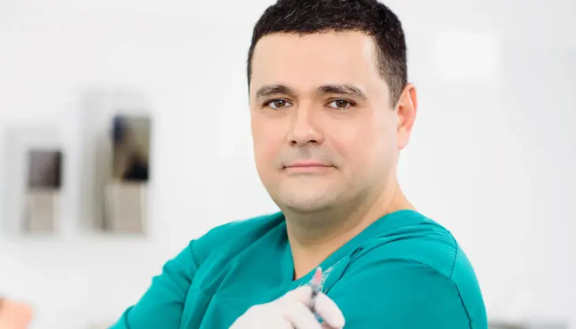 Lekarz w zielonym fartuchu trzyma strzykawkę. W tle widać elementy gabinetu. Wiele pozytywnych klinika ambroziak opinie potwierdza jego profesjonalizm.