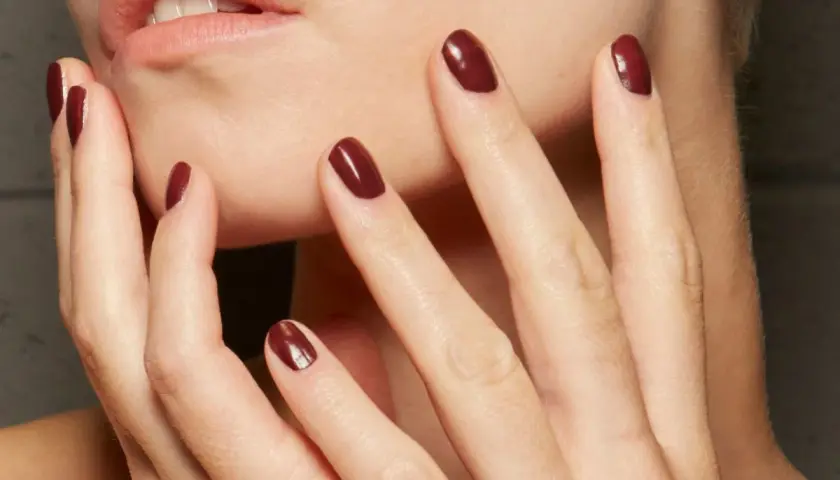 Dłonie z idealnym, ciemnoczerwonym manicurem kombinowanym. Delikatne muśnięcie policzka podkreśla elegancję.