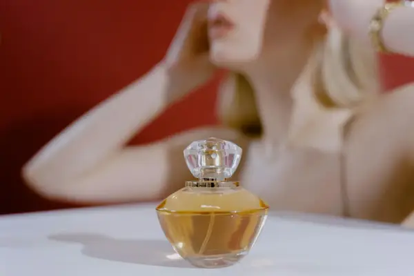 Elegancka buteleczka perfum z płynem o barwie **citral** na białym tle, w tle niewyraźna sylwetka kobiety.